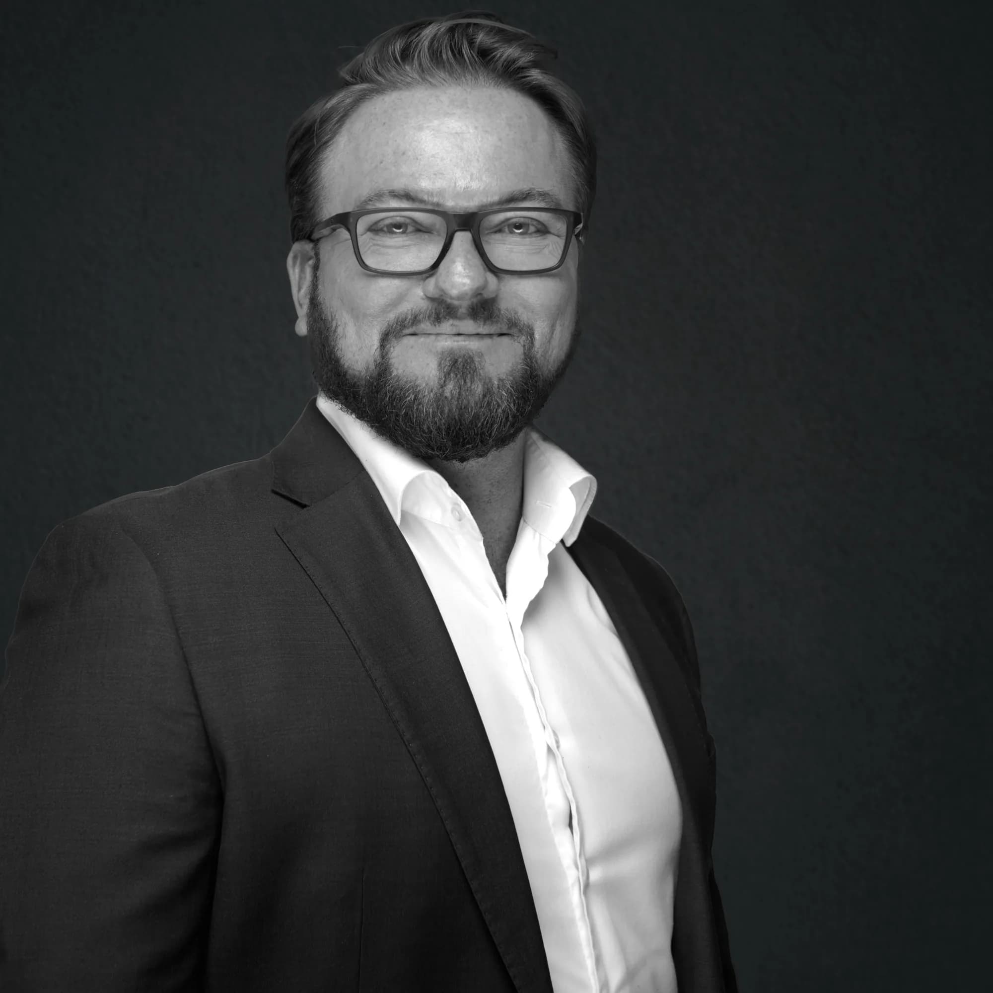 Black-and-white corporate headshot of a bearded man on a dark backdrop